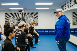 Grandmaster Stephen Del Castillo, founder and lead master instructor of Krav Maga Martial Arts, wearing a blue jacket and cap while speaking to a line of students kneeling on the blue mat in the dojo.