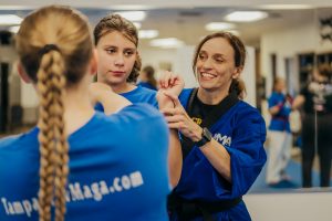 A smiling female Krav Maga instructor in a blue uniform and black belt holds a young student's wrist to guide proper hand and arm positioning during a self-defense class. In the foreground, another student wearing a blue "TampaKravMaga.com" shirt watches closely.