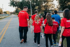 A group of adults and children wearing matching red shirts with "KMMAUSA.COM" on the back walk together away from the camera down a paved suburban street.