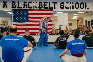 Inside a martial arts dojo, instructor Grandmaster Stephen Del Castillo stands on blue mats in front of a large American flag, gesturing with his hand as students seated on the floor raise their hands to answer a question.