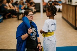 A female martial arts instructor wearing glasses, a blue zip-up jacket, and a black t-shirt kneels down to eye level to talk to a young student wearing a white uniform with a yellow belt. The instructor holds two blue kicking pads in her hand as she speaks kindly to the student, who stands facing her attentively. Parents and spectators sit blurred in the background of the dojo lobby.