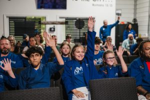 A group of children and adults wearing matching blue Krav Maga Martial Arts (KMMA) uniforms sit in rows of seats during a seminar or class. Several young students in the foreground smile with their hands raised in the air, actively participating.   