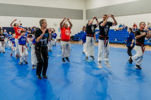 A wide, energetic action shot of martial arts students and instructors practicing together on a large blue mat in a gymnasium. In the center, multiple instructors and adult students mid-air during a jumping exercise, surrounded by children and youth students following along. Blue bleachers line the background wall.