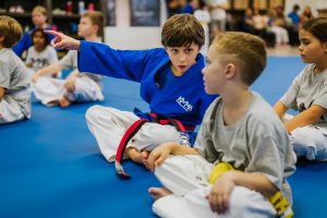 A young martial arts student in a blue KMMA gi sits on a blue mat and points across the room while talking to a fellow student in a gray shirt.