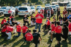 An outdoor martial arts gathering on a grassy field surrounded by parked vehicles on a sunny day. Dozens of participants—children, teens, and adults—are arranged in a large circle kneeling on one knee or sitting in listening poses. Many wear red or black t-shirts printed with "KMMAUSA.COM". At the center of the circle, Grandmaster Del Castillo is in a red t-shirt, baseball cap, and sunglasses gesturing with both hands as he addresses the group.