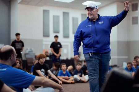 At Krav Camp, Grandmaster Del Castillo walks the floor and his left arm is raised. There are students sitting all around.